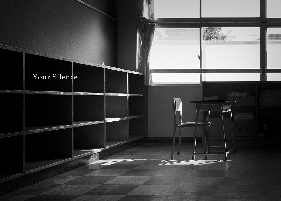 モノクロ写真、小学校の教室の中。ひとつの机と椅子。その後ろに棚がある。