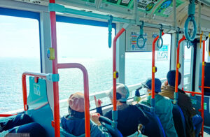 バスの中、窓から海が見える。外を見て指さしながら会話する女性たち。